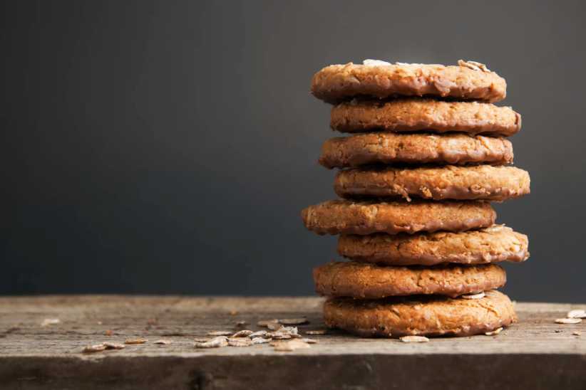 Stack of homemade oatmeal cookies on rustic wooden surface with scattered oats, against a dark gray background.