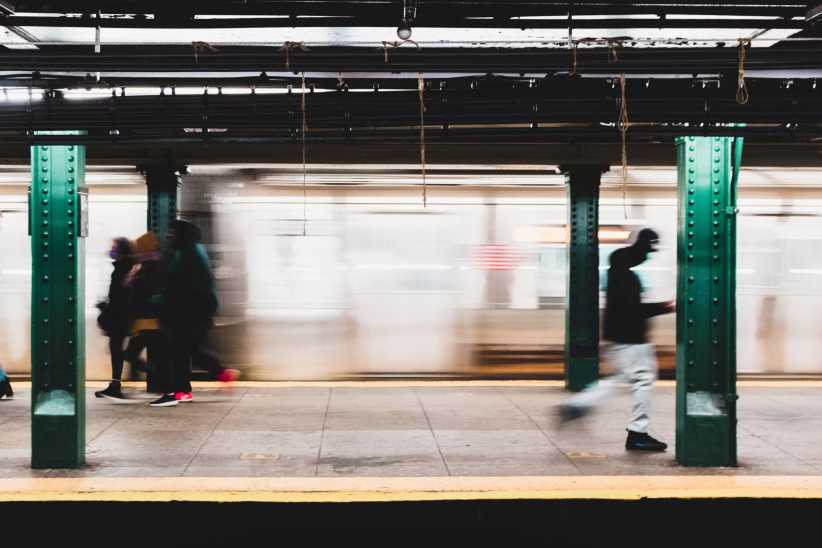 People walking and waiting on a subway platform with green columns, while a train speeds by in the background, creating a motion blur effect.