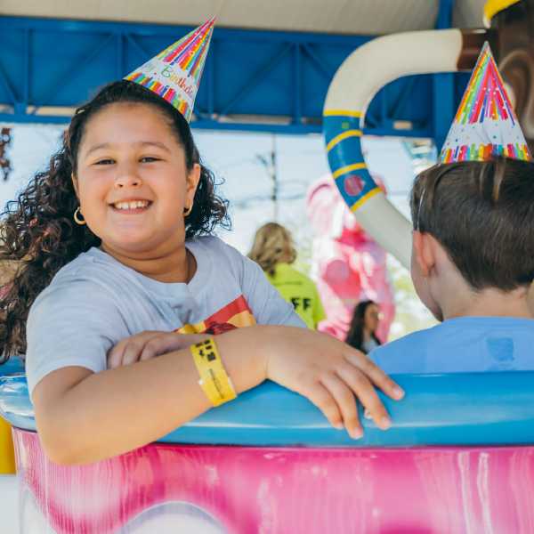Two children wearing birthday hats sit together on a colorful amusement park ride, enjoying a festive celebration outdoors.