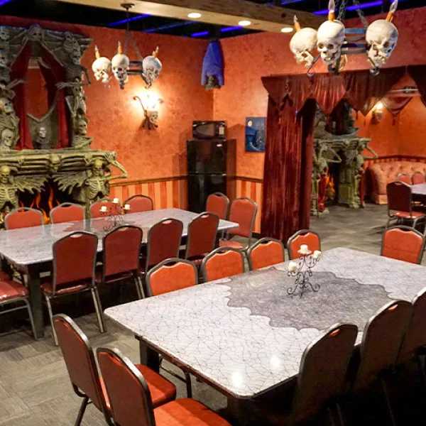 A themed dining room features spiderweb-like tables, red chairs, skull chandeliers, and gothic decor with skeleton fireplace surrounds. A themed dining room features spiderweb-patterned tables, red chairs, skull chandeliers, and gothic skeleton decorations throughout.