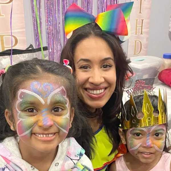 A woman with a rainbow bow smiles with two girls, both with colorful butterfly face paint, one wearing a crown, at a festive party.