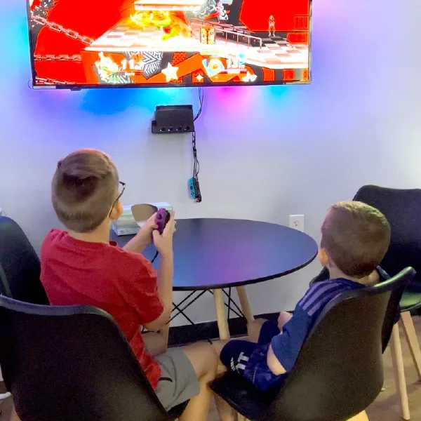 Two boys sit at a round table playing a colorful video game on a wall-mounted TV in a cozy, well-lit gaming room.