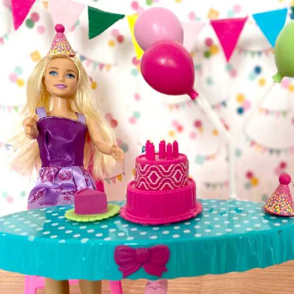 A blonde doll wearing a party hat and purple dress sits at a table with a pink birthday cake, balloons, and festive decorations.
