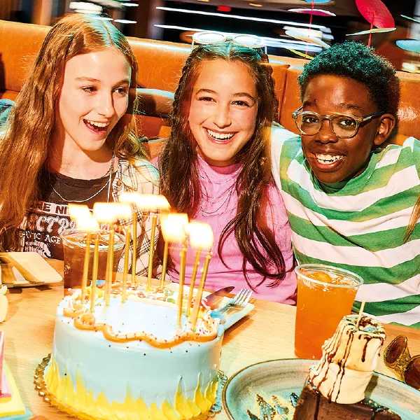 Three kids sit closely together at a restaurant booth, celebrating with a birthday cake, drinks, and dessert in front of them.