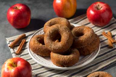 A plate of cinnamon sugar-coated apple cider doughnuts sits on a striped cloth, surrounded by fresh apples and cinnamon sticks.