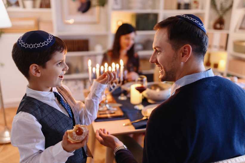 Smiling father and son celebrating Hanukkah, with a lit menorah, festive dinner table, and a jelly doughnut in the boy’s hand.