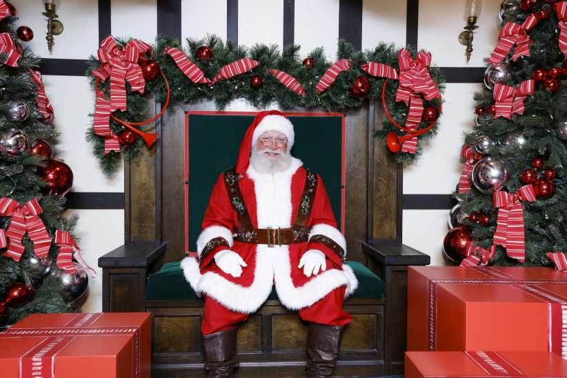 Santa Claus sits smiling on a festive chair, surrounded by holiday garlands, red bows, ornaments, and large red gift boxes.