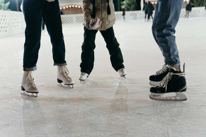 Three people wearing ice skates stand on an outdoor rink, dressed warmly and preparing to skate on the smooth ice surface.Three people wearing ice skates stand on an outdoor rink, dressed warmly and preparing to skate on the smooth ice surface.