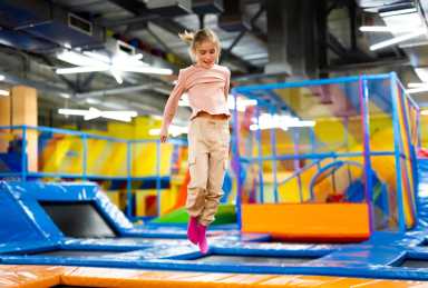 A young girl in pink socks and beige pants jumps joyfully on a trampoline at a colorful indoor play park.