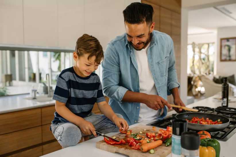 A father and son happily cook together in a modern kitchen, chopping vegetables and preparing a colorful meal on the counter.