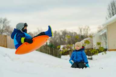 Child sledding down a snowy hill using an orange disc while another child in a blue coat watches beside houses and trees.