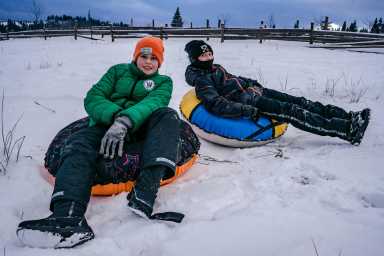 Two people wearing winter clothing sit on snow tubes in a snowy field, relaxing after sledding with a wooden fence in the background.