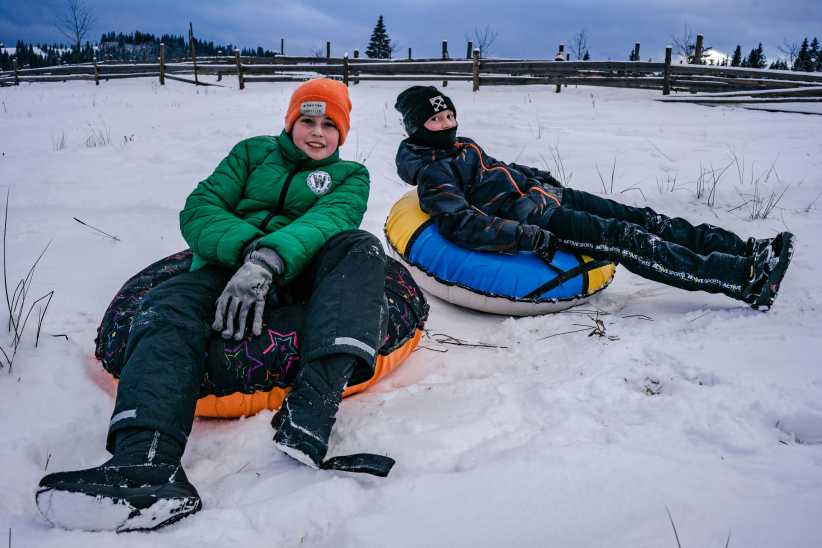 Two people wearing winter clothing sit on snow tubes in a snowy field, relaxing after sledding with a wooden fence in the background.