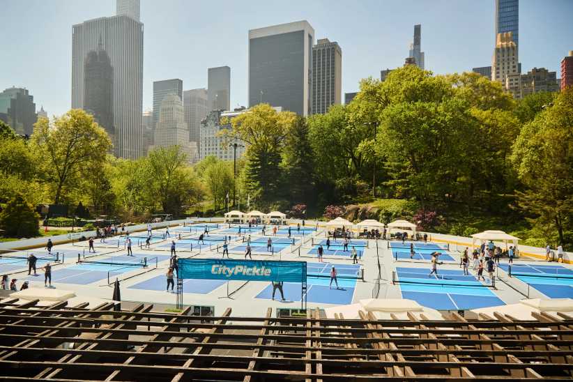 Multiple blue pickleball courts in Central Park’s Wollman Rink with skyscrapers and green trees in the background, people playing.