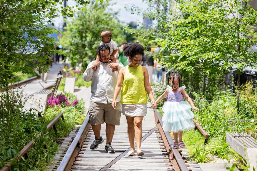 A family of four, including a toddler and girl in a blue dress, walks along the lush green High Line park in New York City.
