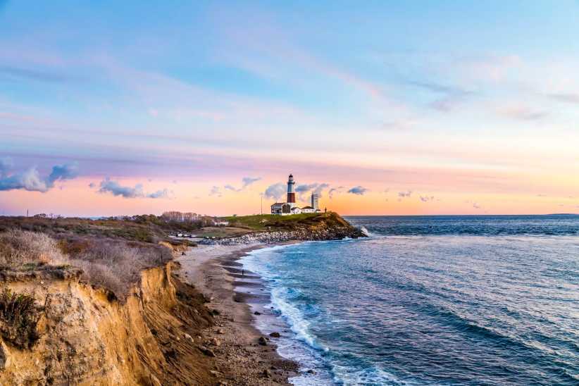 A lighthouse stands on a grassy cliff at sunset, overlooking a rocky shoreline and calm ocean under a pastel sky.