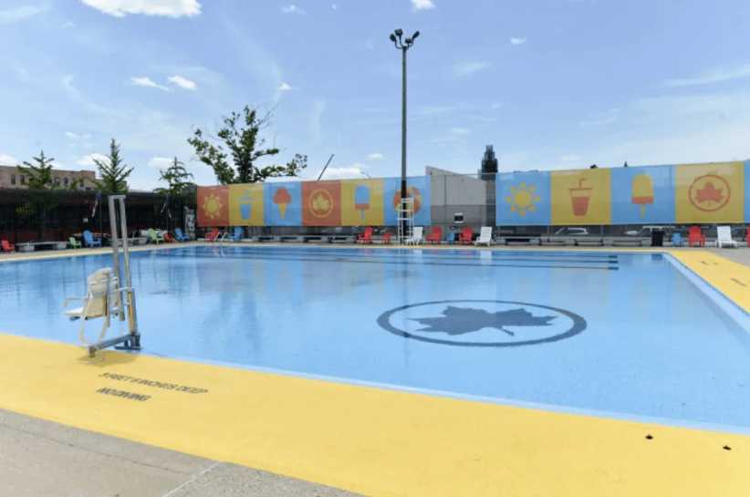 A clear, outdoor swimming pool with a blue maple leaf at the center and colorful wall murals, surrounded by yellow pavement.