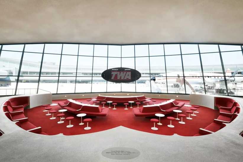 A retro-style airport lounge with red seating and white tables, large windows overlook planes on the runway outside.