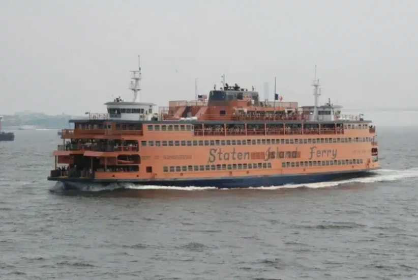 An orange Staten Island Ferry sails across the water, carrying passengers on multiple decks under a cloudy sky.