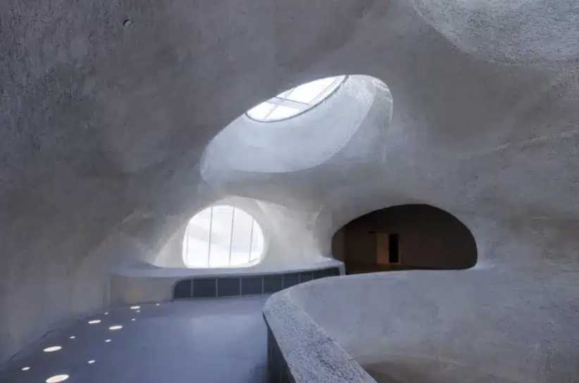 A smooth, organic interior space with curved white walls and large skylights letting in natural light, resembling a cave or dome.