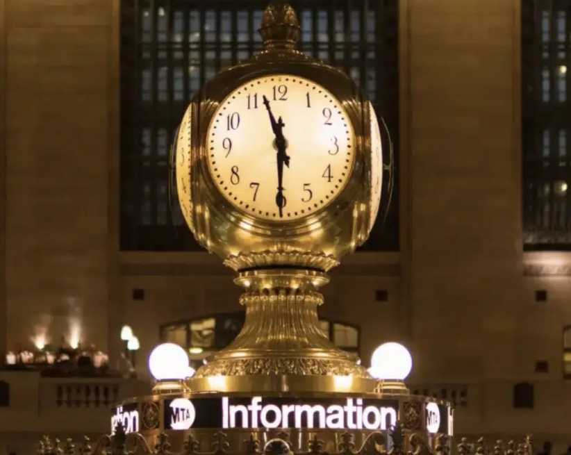A classic brass clock sits atop an information booth, glowing warmly inside a grand hall with tall windows in the background.