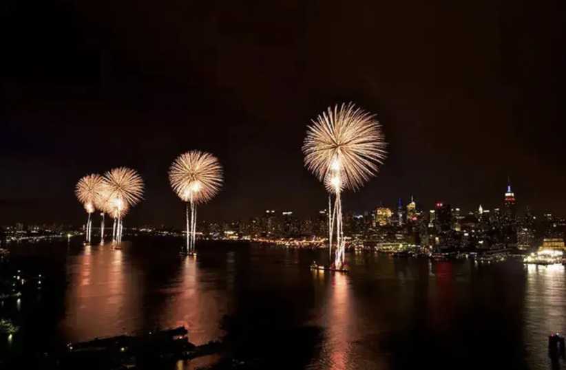 Fireworks burst in the night sky over a city skyline, illuminating the water below and creating colorful reflections on the surface.
