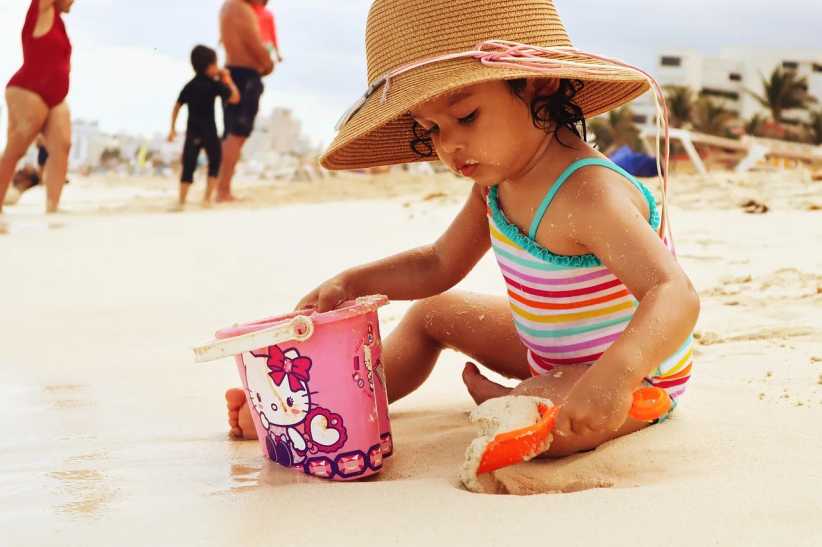 A young child in a striped swimsuit and sunhat plays with a shovel and Hello Kitty bucket in the sand on a lively beach.