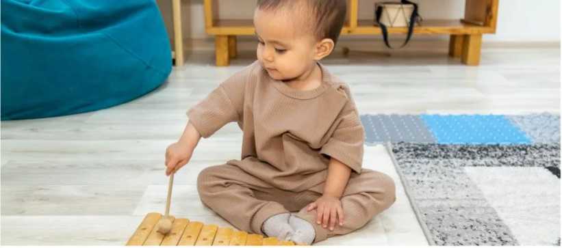 A toddler in neutral-colored clothes sits on the floor playing a wooden xylophone with a mallet in a brightly lit indoor room.