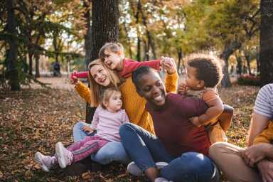 A group of adults and children sit together on the ground in a park during autumn, surrounded by trees and fallen leaves.