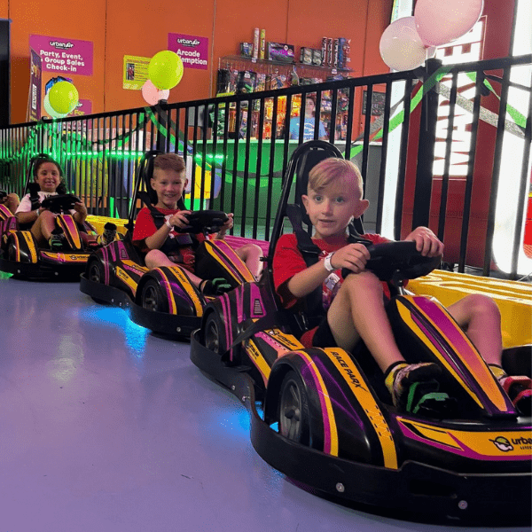 Children sit in a row of colorful mini go-karts indoors, ready to race, with balloons and party decorations in the background.