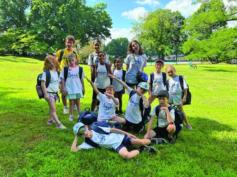 A group of smiling kids and camp counselors wearing "Oasis Day Camp" shirts pose together on a sunny day in a large green field.