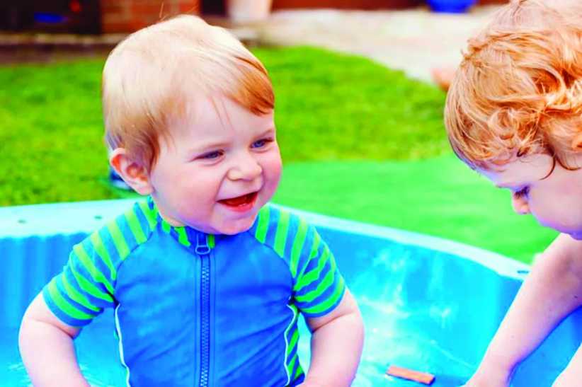 Two toddlers play happily in a small blue kiddie pool outdoors, smiling and splashing water on a sunny day in the backyard.