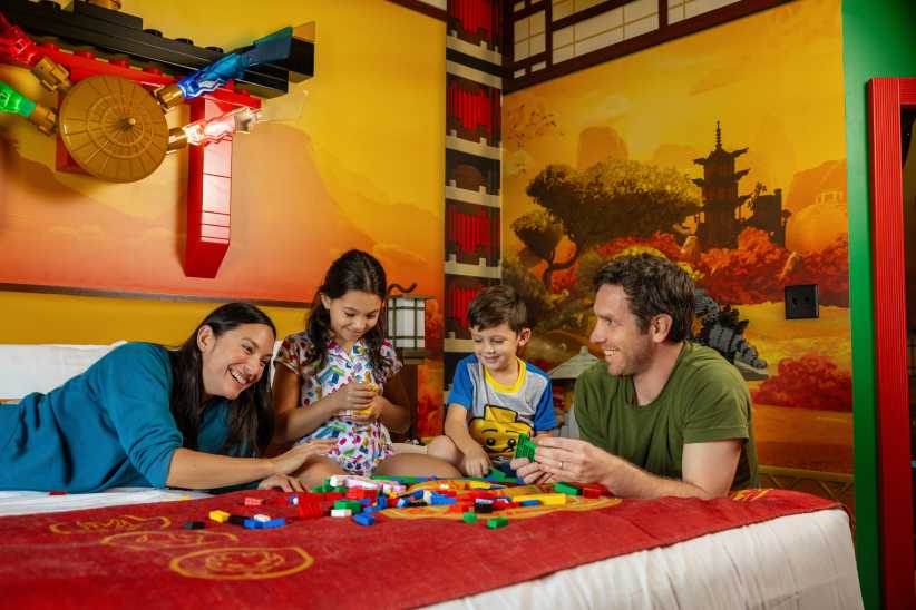 A family sits together on a bed playing with colorful LEGO bricks in a cozy, Asian-themed room decorated with pagoda wall art.