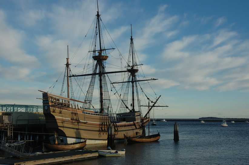 A historic wooden sailing ship with tall masts and rigging is docked by the shore, with small boats and calm water under a blue sky.