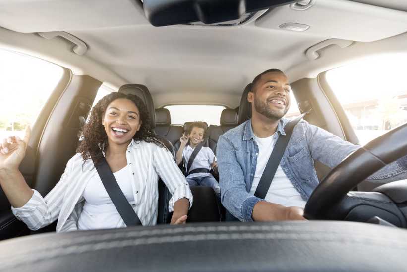 A family sits inside a car with seatbelts fastened; the man drives while the woman gestures and the child relaxes in the back seat.