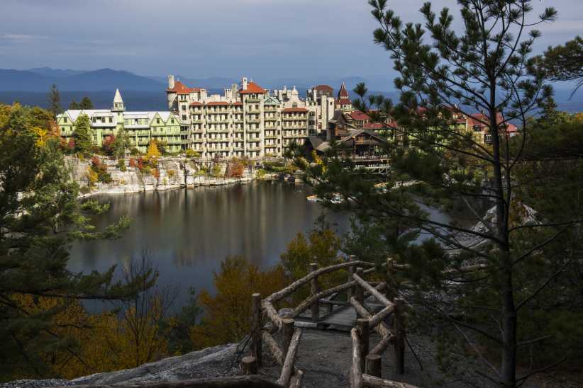 Grand lakeside resort with castle-like architecture stands beside a tranquil water body, surrounded by forest and a rustic wooden railing in front. Large resort buildings with castle-like towers overlook a serene lake, surrounded by lush forest and a rustic wooden fence in the foreground.