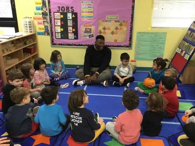 A teacher sits cross-legged on the floor in a circle with young children in a colorful preschool classroom, engaging in group time.