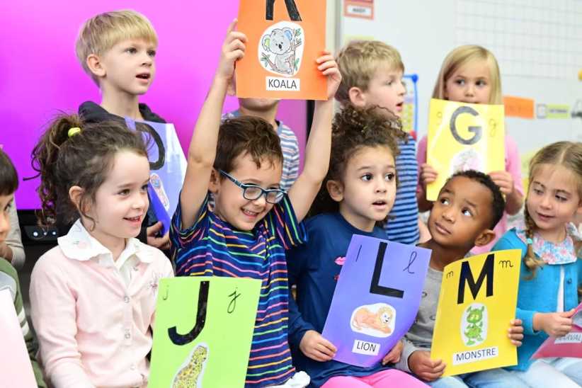 A group of young children in a classroom hold up colorful cards with letters and animal illustrations, participating in an alphabet activity.