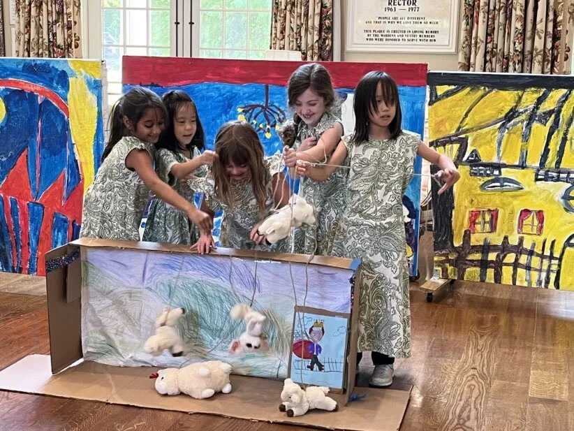 Five young girls manipulate marionettes of lambs in a makeshift puppet theater constructed from cardboard. The backdrop features child-like drawings. The scene takes place in a room with hardwood floors and large, colorful paintings.