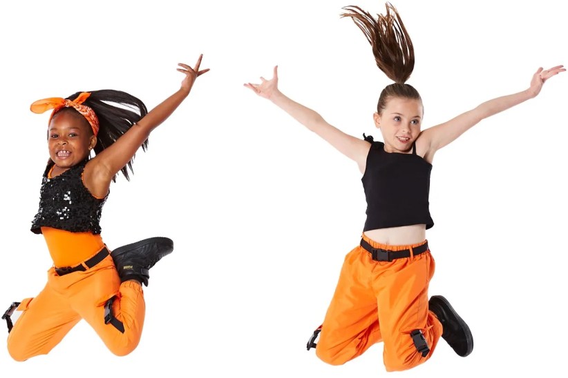 Two girls, wearing orange pants and tops, joyfully leap against a white background. One girl wears a black sequined top and a headband.