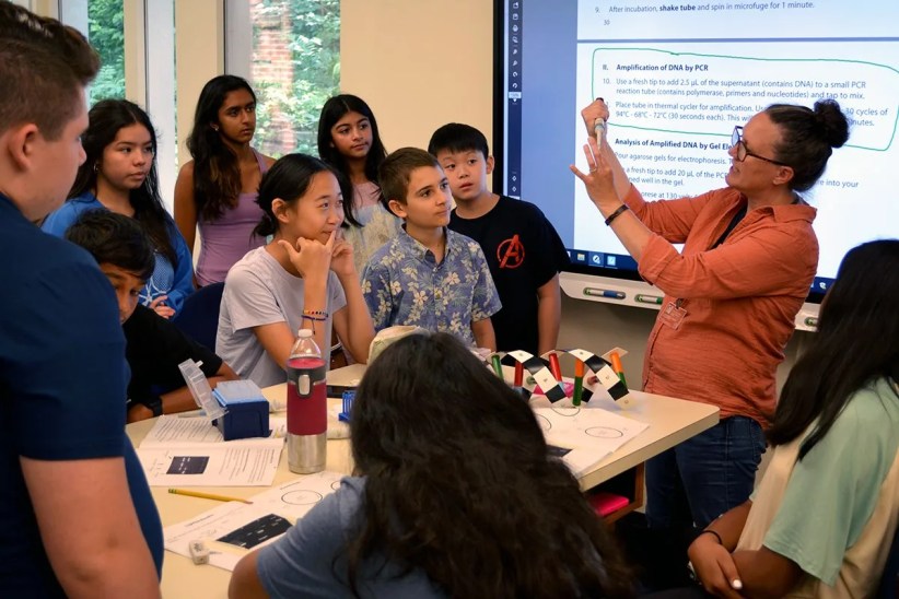 A teacher demonstrates a lab procedure, using a pipette to explain steps to a group of attentive middle school students gathered around a table in a classroom.