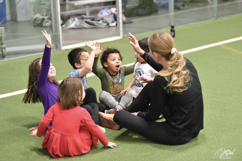 Young children and a woman are sitting on a green indoor playing field. They are playing a game involving raised hands. One child's shirt says "Mighty Toys Soccer".