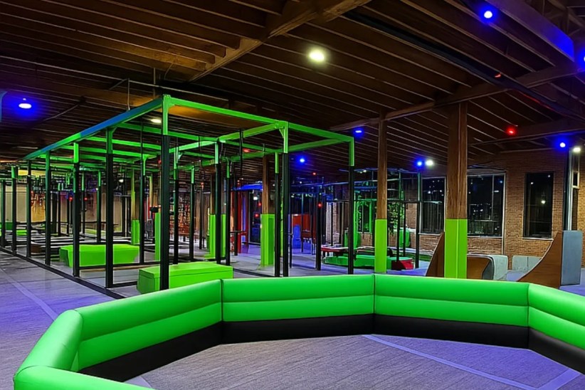 An indoor obstacle course, brightly lit in green and blue, is situated within a large warehouse-like space with exposed wooden beams and brick walls. A large, padded green seating area is in the foreground.