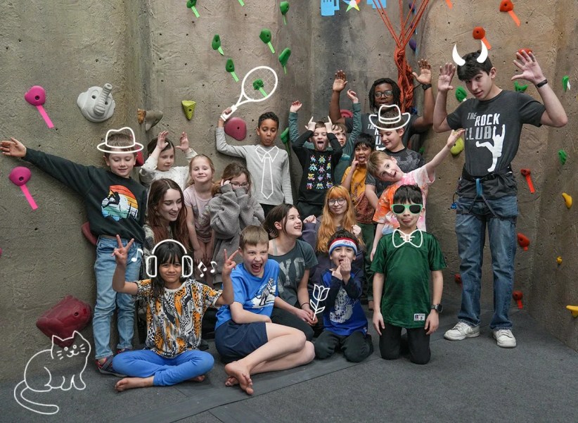 A large group of children and a young adult pose playfully near an indoor climbing wall. Many make silly faces; some have drawn-on accessories. One shirt reads, "THE ROCK CLUB."