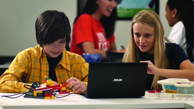 Two children construct a Lego robot at a table using a laptop; other students and a teacher are visible in the background of a classroom. The laptop's brand is "ASUS".