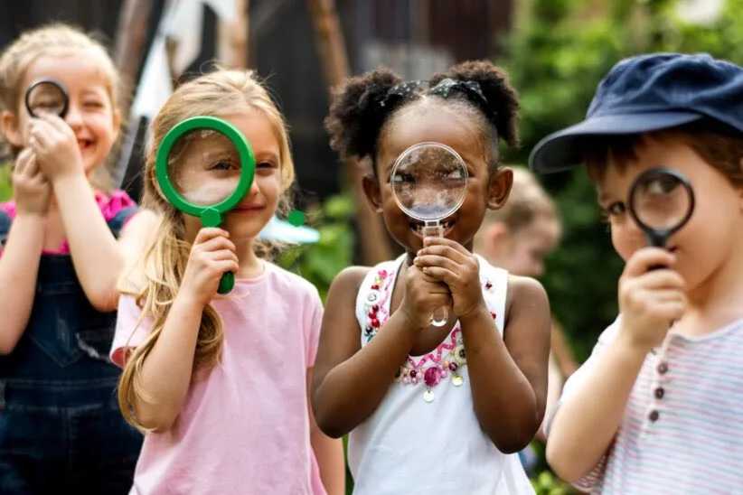 Young children hold magnifying glasses to their eyes, appearing to explore their surroundings in an outdoor setting.