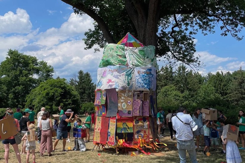 A large, colorful cardboard structure, decorated with painted fabric and children’s artwork, stands in a grassy park under a large tree. People, some carrying cardboard boxes, are gathered around it.