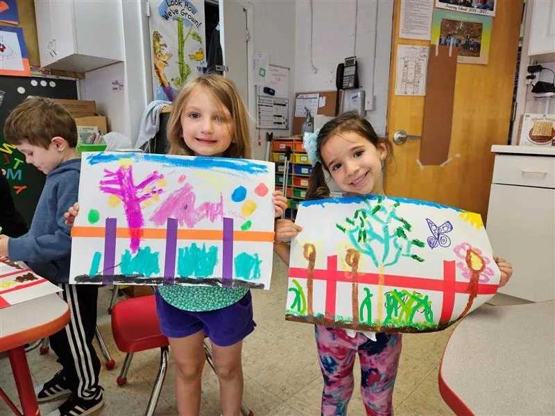 Two young children proudly display their colorful paintings in a classroom, smiling as they hold their artwork up for the camera.