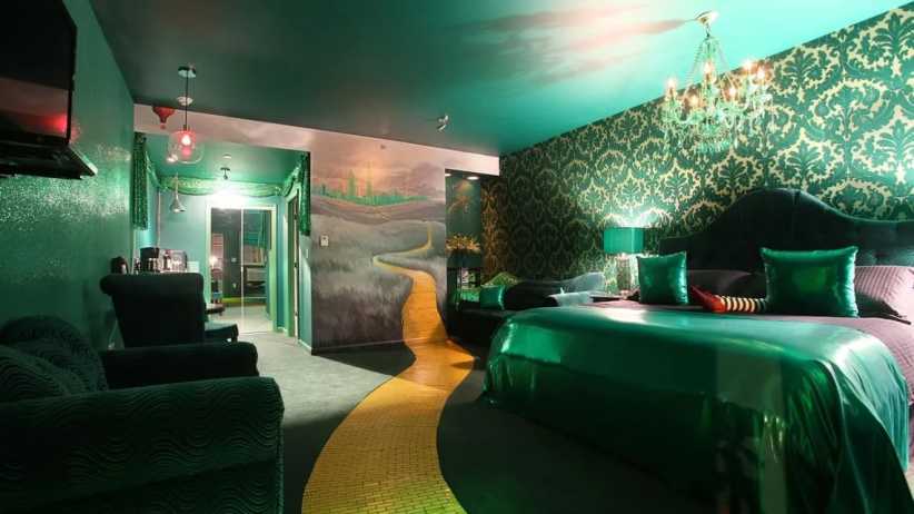 A whimsical, emerald-themed bedroom features ornate green wallpaper, sparkling chandelier, cozy seating, and a yellow brick road design on the floor leading to a mural of a fantasy landscape.