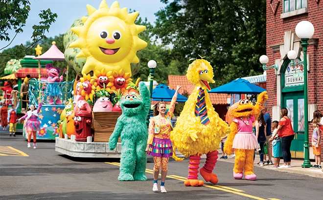 Sesame Street parade with Big Bird, Oscar the Grouch, and other costumed characters marching past colorful floats and cheering crowds.